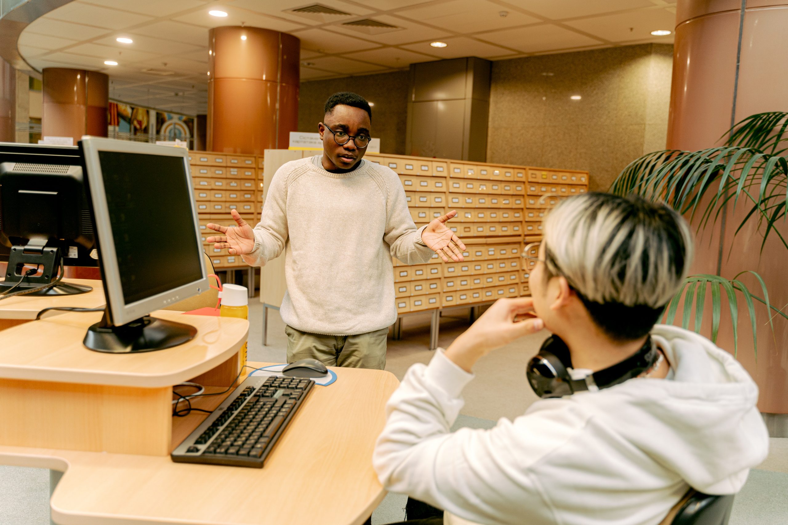 A young male adult gesticulates to another young male adult, who is sitting at a computer desk in an office space.